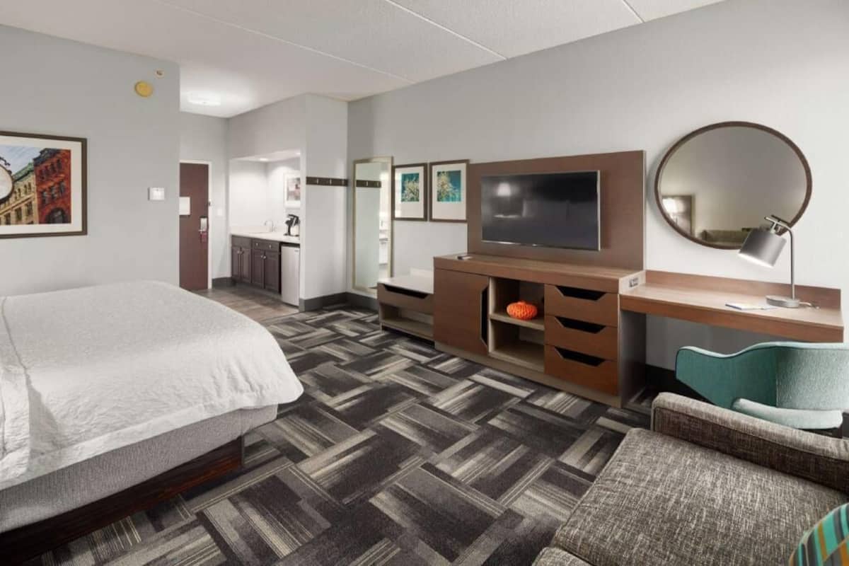 A hotel room with a bed, a TV, a desk, a chair, a mirror, and a carpeted floor.
