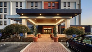 Une entrée d’hôtel moderne, dotée d’une allée couverte, de plantes en pot et d’un panneau indiquant un espace de stationnement.