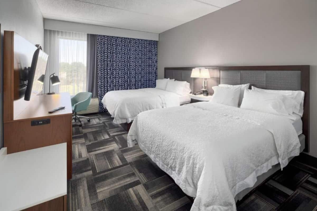 A hotel room with two beds, a desk, a chair, and a large window with curtains.