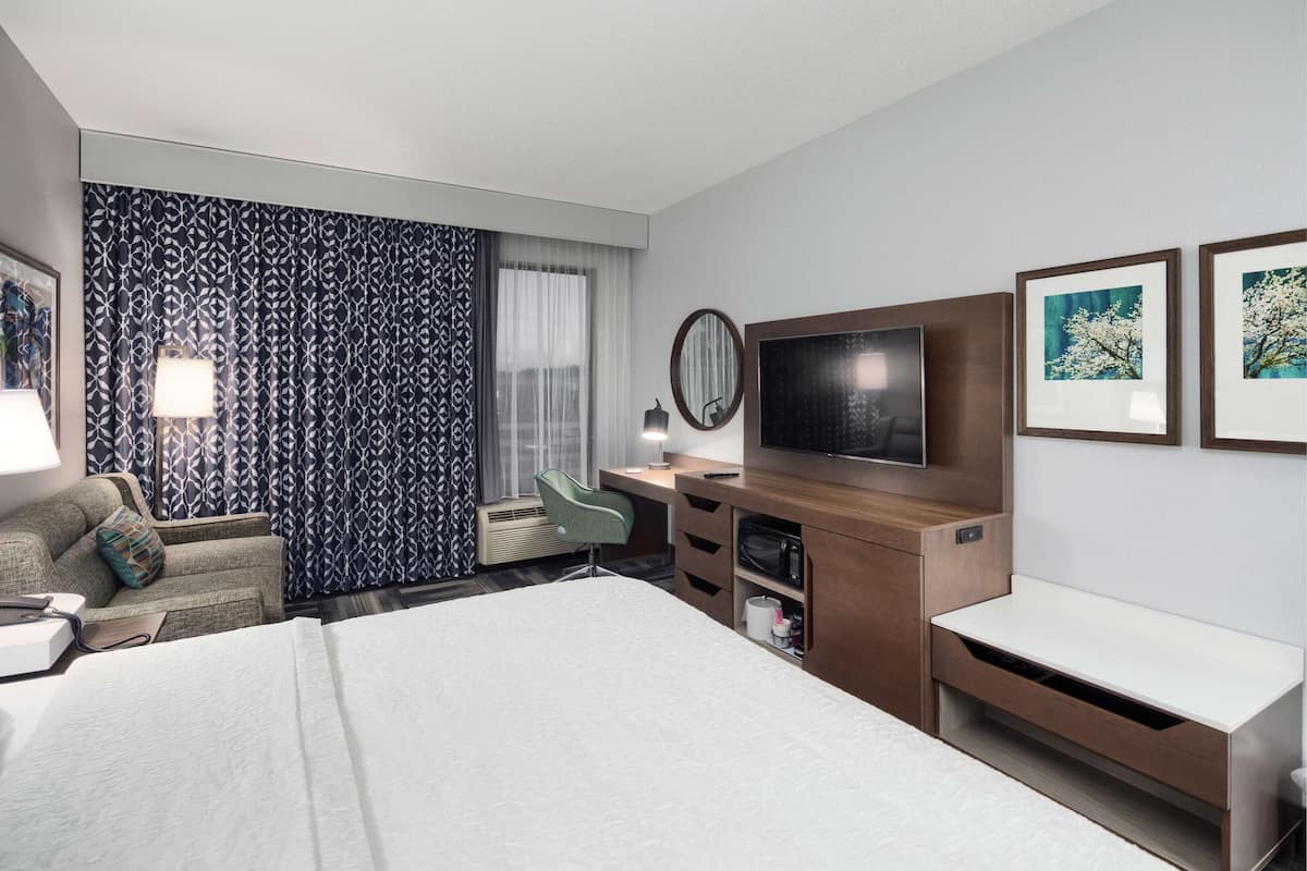 A hotel room with a large bed, a desk with a flat-screen TV, a chair, and a large window with patterned curtains.