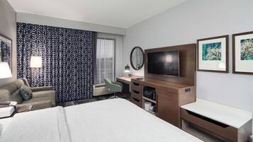 Habitación de hotel con una cama grande, un escritorio con televisor de pantalla plana, una silla y un ventanal con cortinas estampadas.