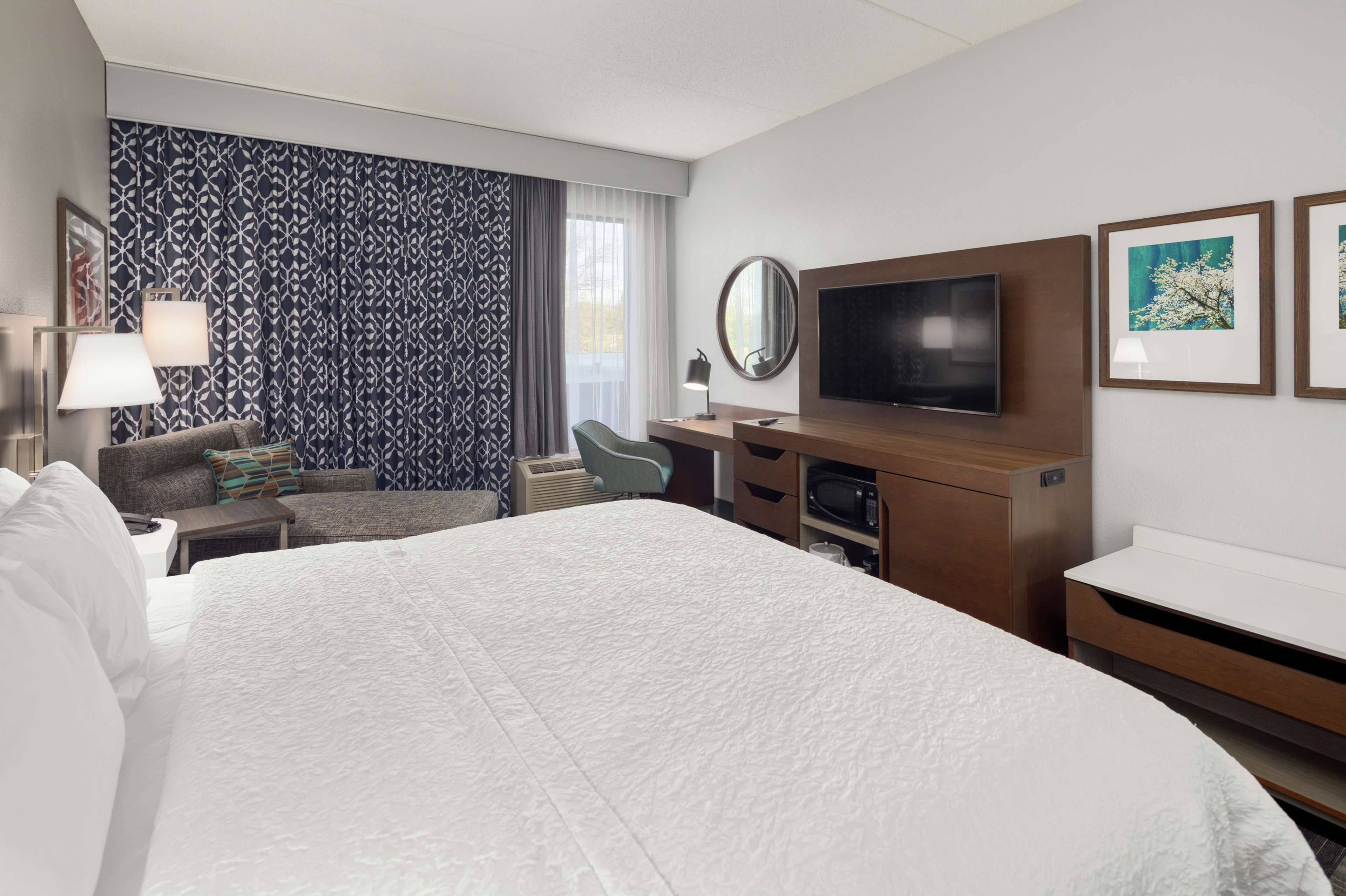 Una habitación de hotel con una cama grande, un escritorio con televisor de pantalla plana, una silla, una lámpara de escritorio y un ventanal con cortinas estampadas.