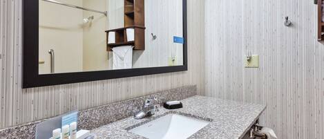 A bathroom with a granite countertop, a white sink, a large mirror, and a wooden shelf with towels.