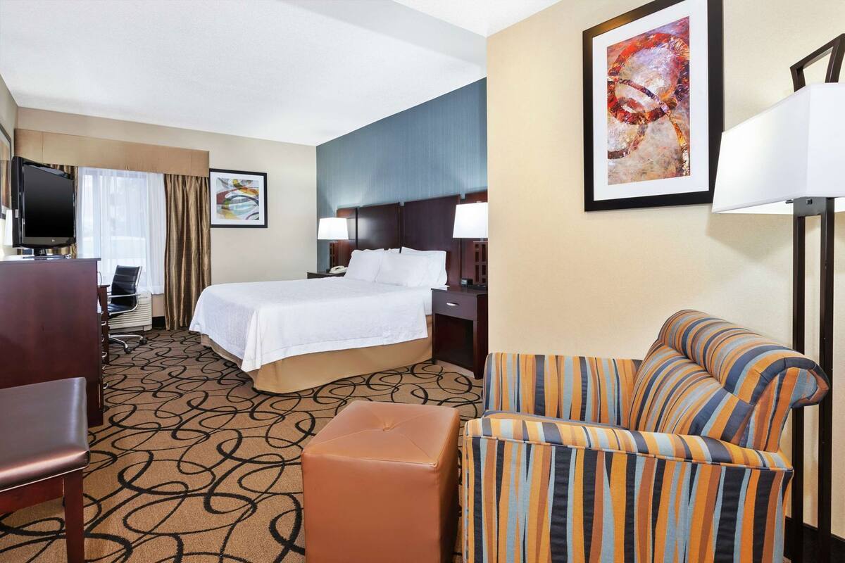 A hotel room with a bed, a sofa, a TV, a desk, and a painting on the wall.