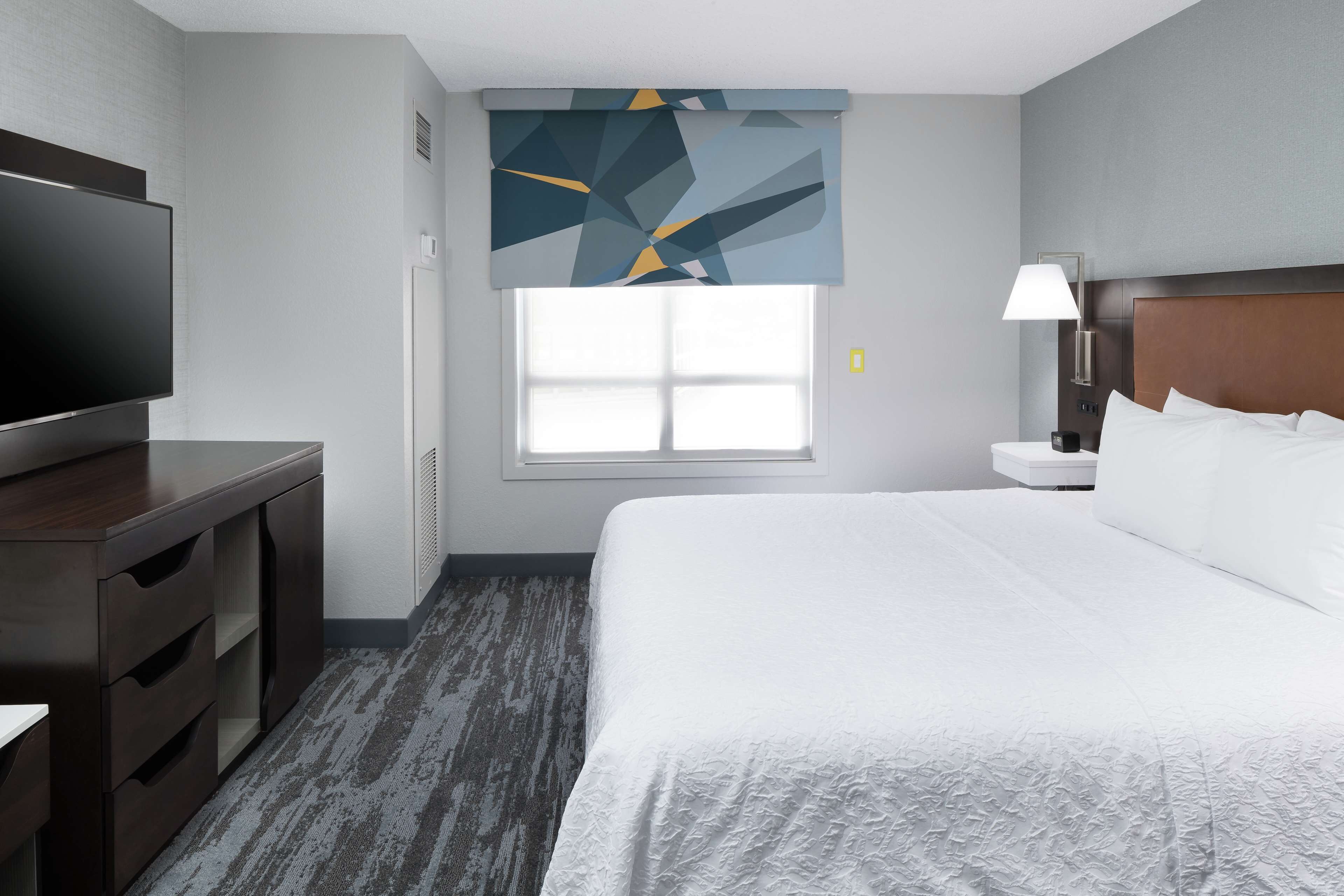 A hotel room with a large bed, a flat-screen TV, a desk with drawers, a window with blinds, and a wall-mounted lamp.