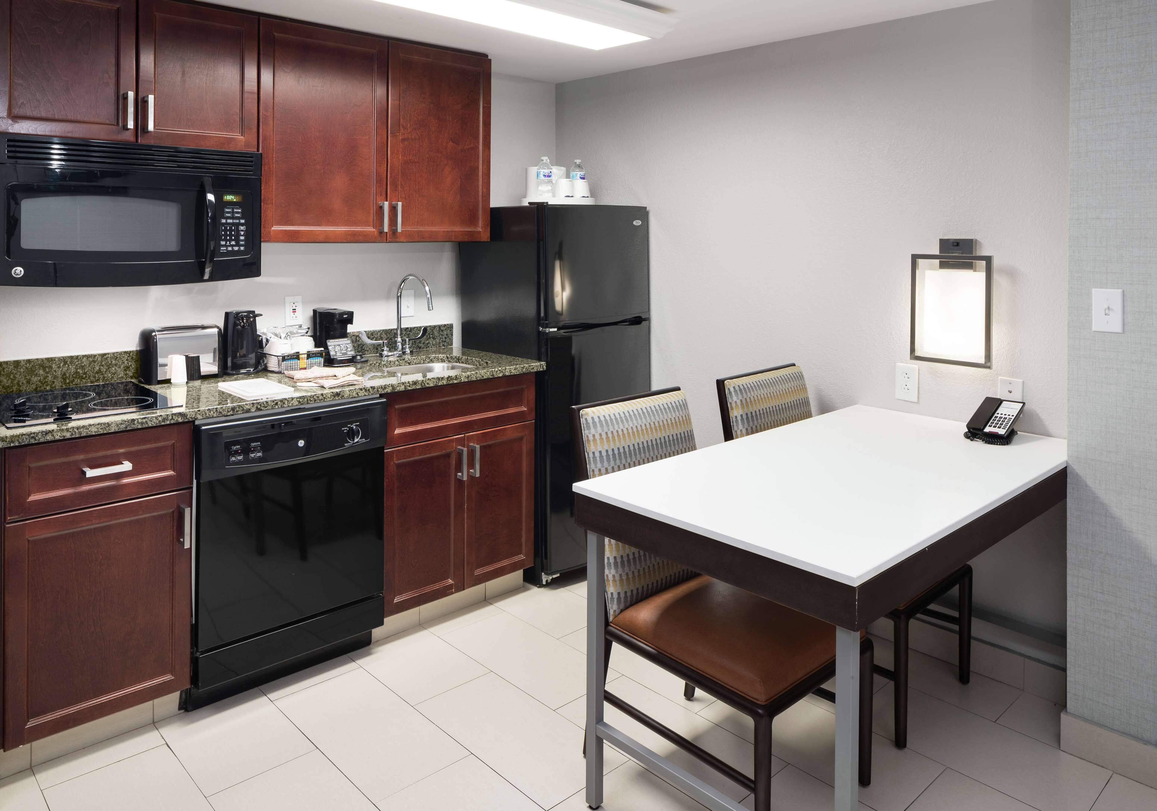 A compact kitchen with dark wood cabinets, a black microwave, a black refrigerator, a coffee maker, and a white table with a chair.
