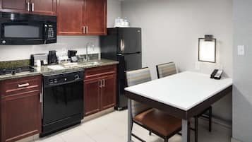 Une cuisine de taille réduite avec des meubles de couleur bois foncé, un micro-ondes noir, un réfrigérateur noir, une machine à café et une table blanche avec une chaise.