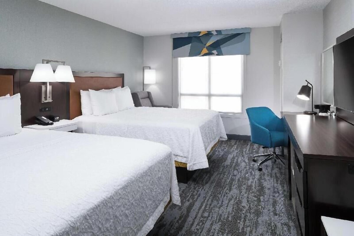 A hotel room with two beds, a desk, a chair, a TV, and a window.