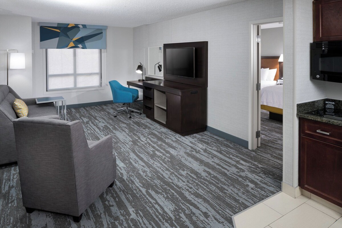 A hotel room with a grey sofa, a blue chair, a desk with a flat-screen TV, a minibar, and a bed in the background.