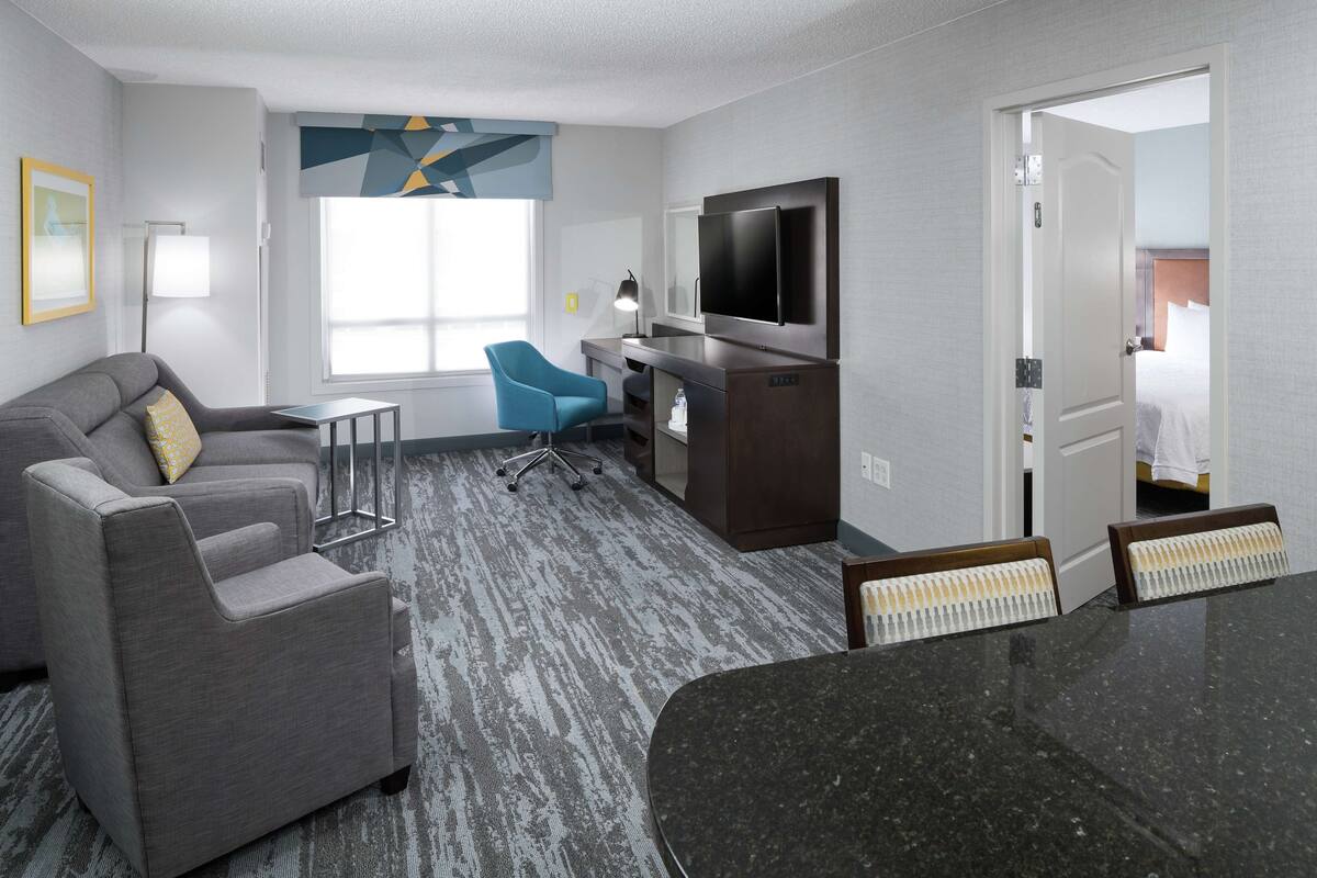 Habitación de hotel moderna con un sofá gris, un sillón azul, una pequeña mesa, un televisor y un baño visible a través de una puerta abierta.