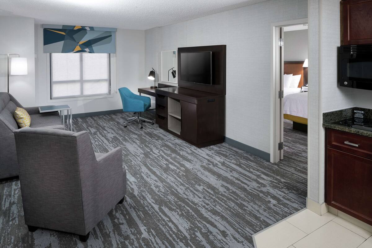 Habitación de hotel con sofá gris, escritorio con televisor de pantalla plana, una silla, una mesita y una pequeña cocina.