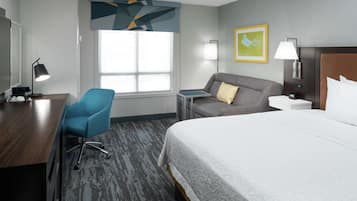 A hotel room with a large bed, a desk, a blue chair, a grey sofa, and a window with a view.