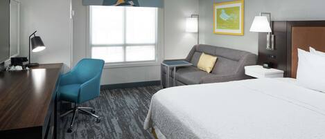 A hotel room with a large bed, a desk, a blue chair, a grey sofa, and a window with a view.