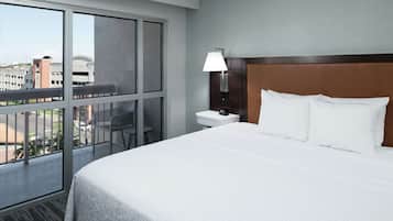 A hotel room with a large bed, a nightstand with a lamp, a chair, and a view of the city through a large window.