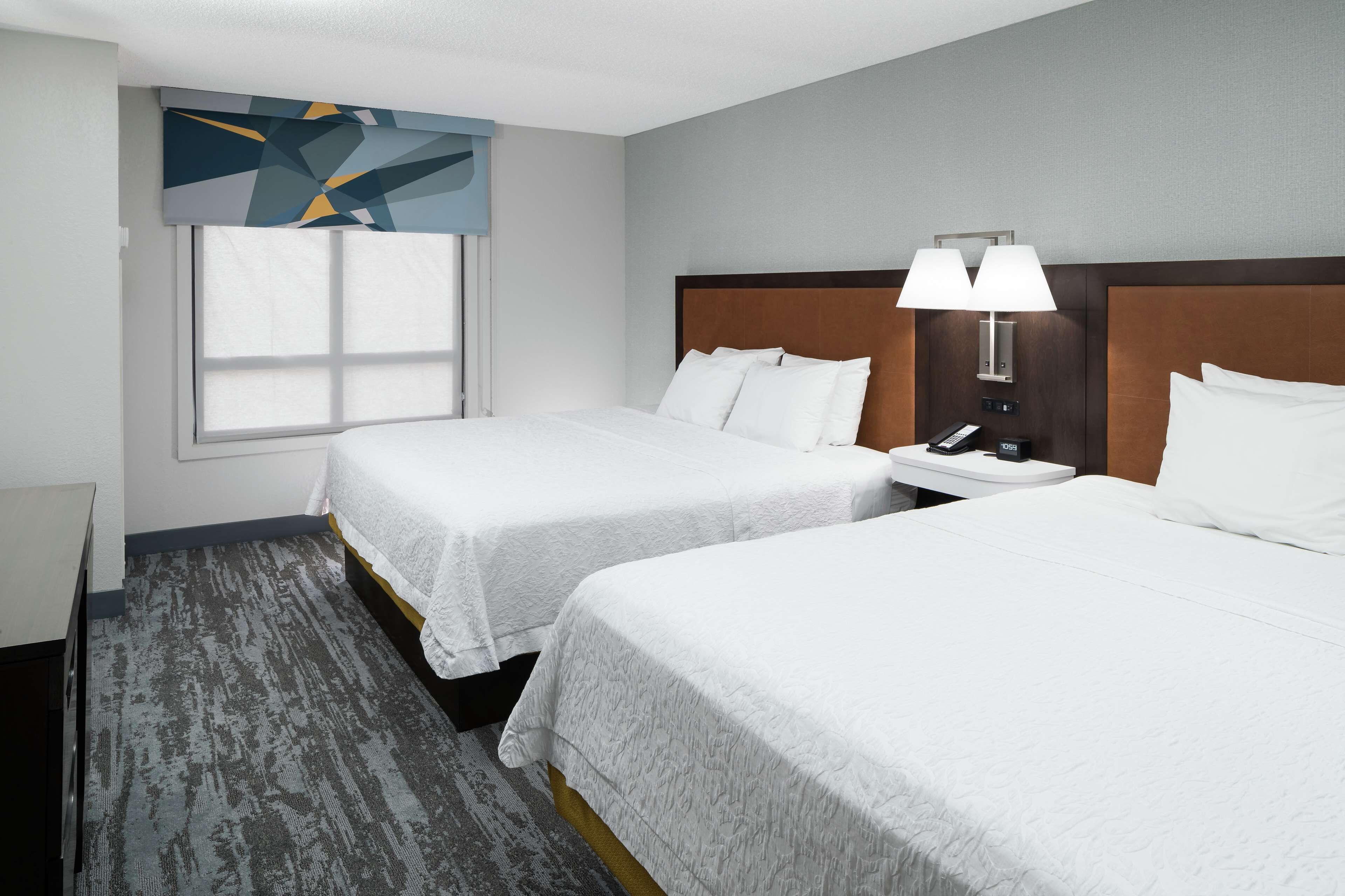 A hotel room with two beds, a nightstand with a lamp, and a window with blinds.