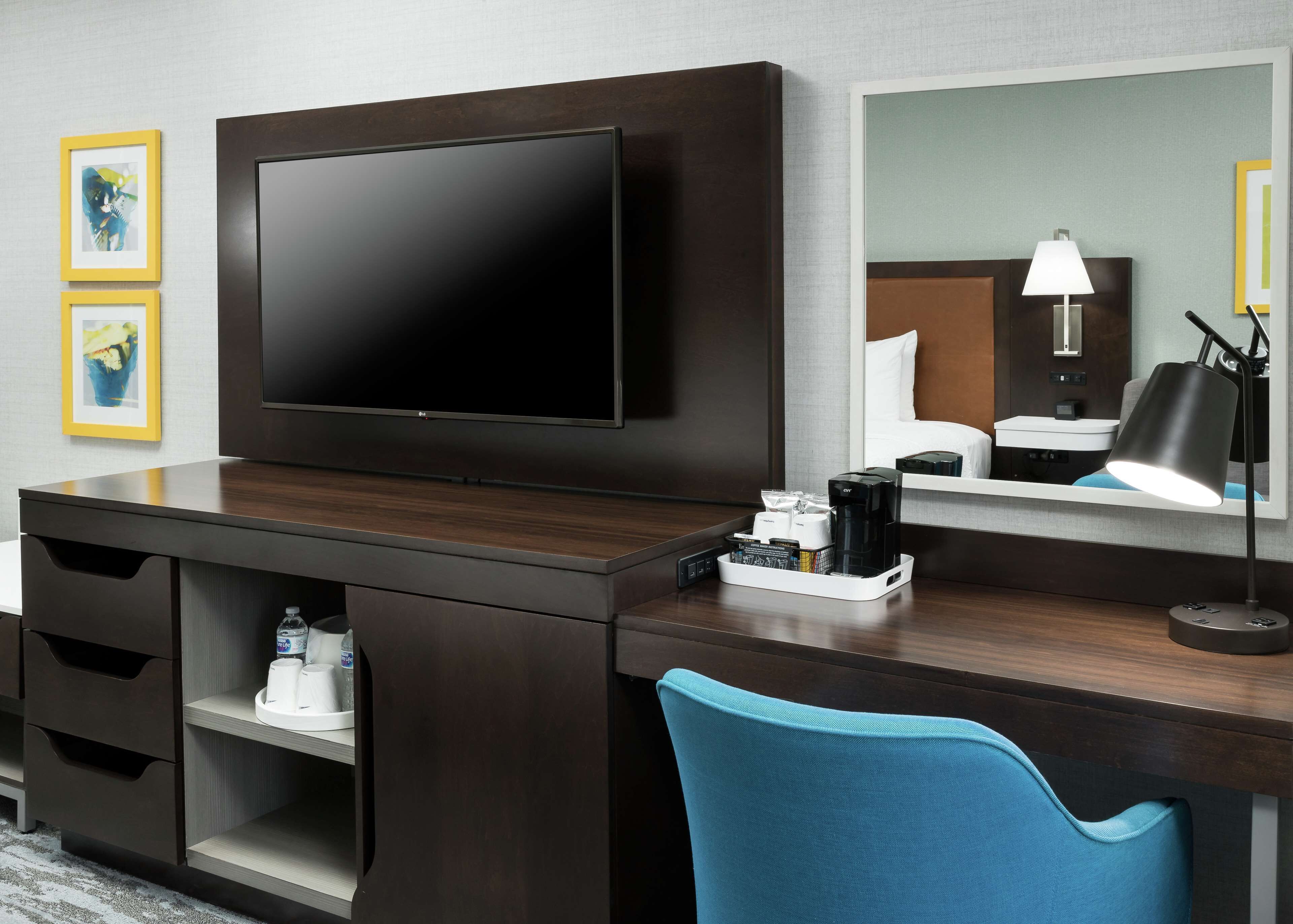 A hotel room with a large flat-screen TV mounted on the wall, a wooden desk with a coffee maker, a lamp, and a chair.
