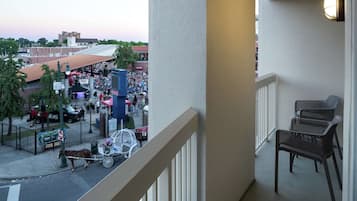 A balcony with wicker chairs, overlooking a busy street with a crowd and a horse-drawn carriage.