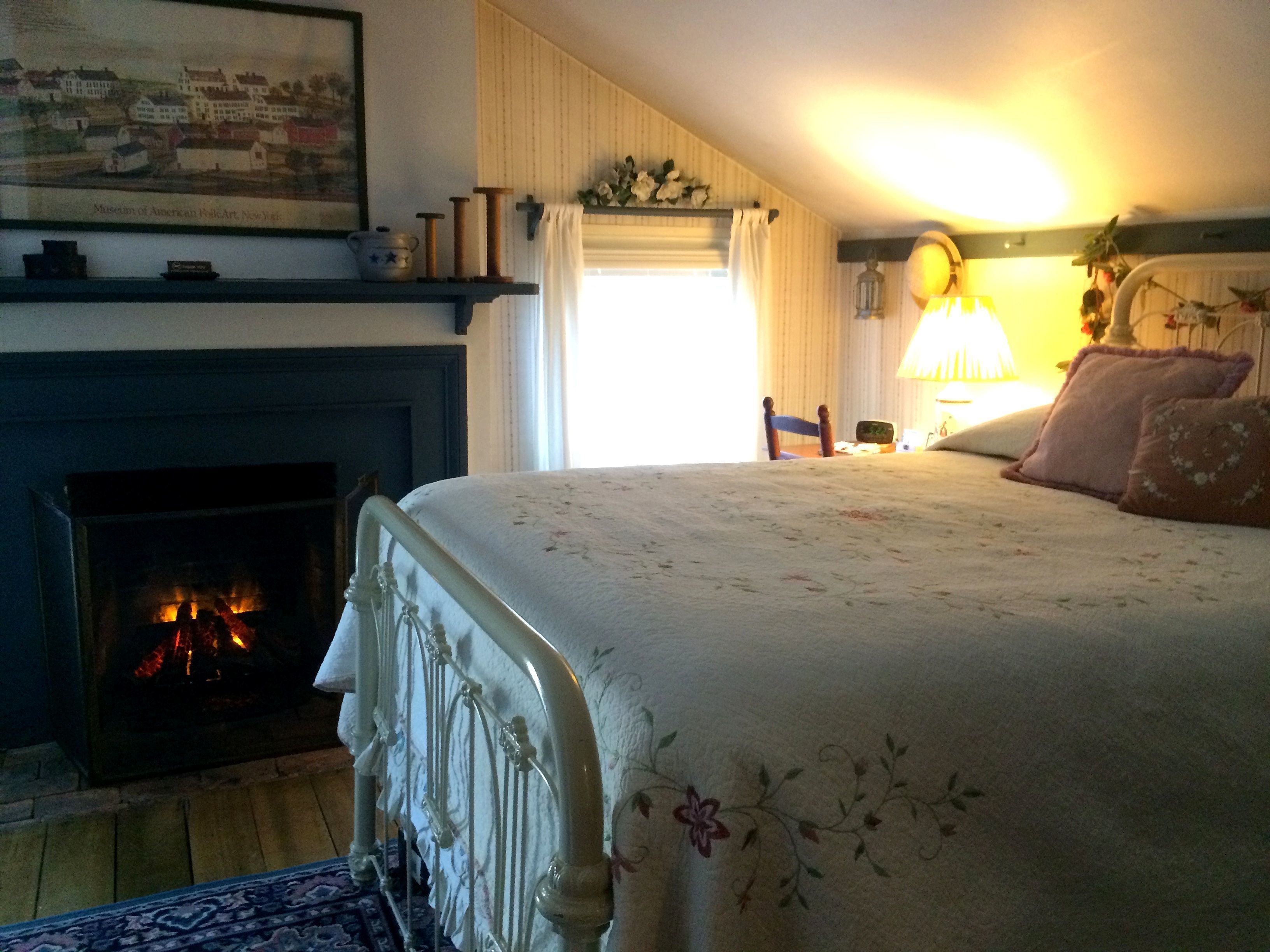 Une chambre avec une cheminée, un lit recouvert d’une literie à motifs floraux, une table de chevet avec une lampe et un tableau accroché au mur.