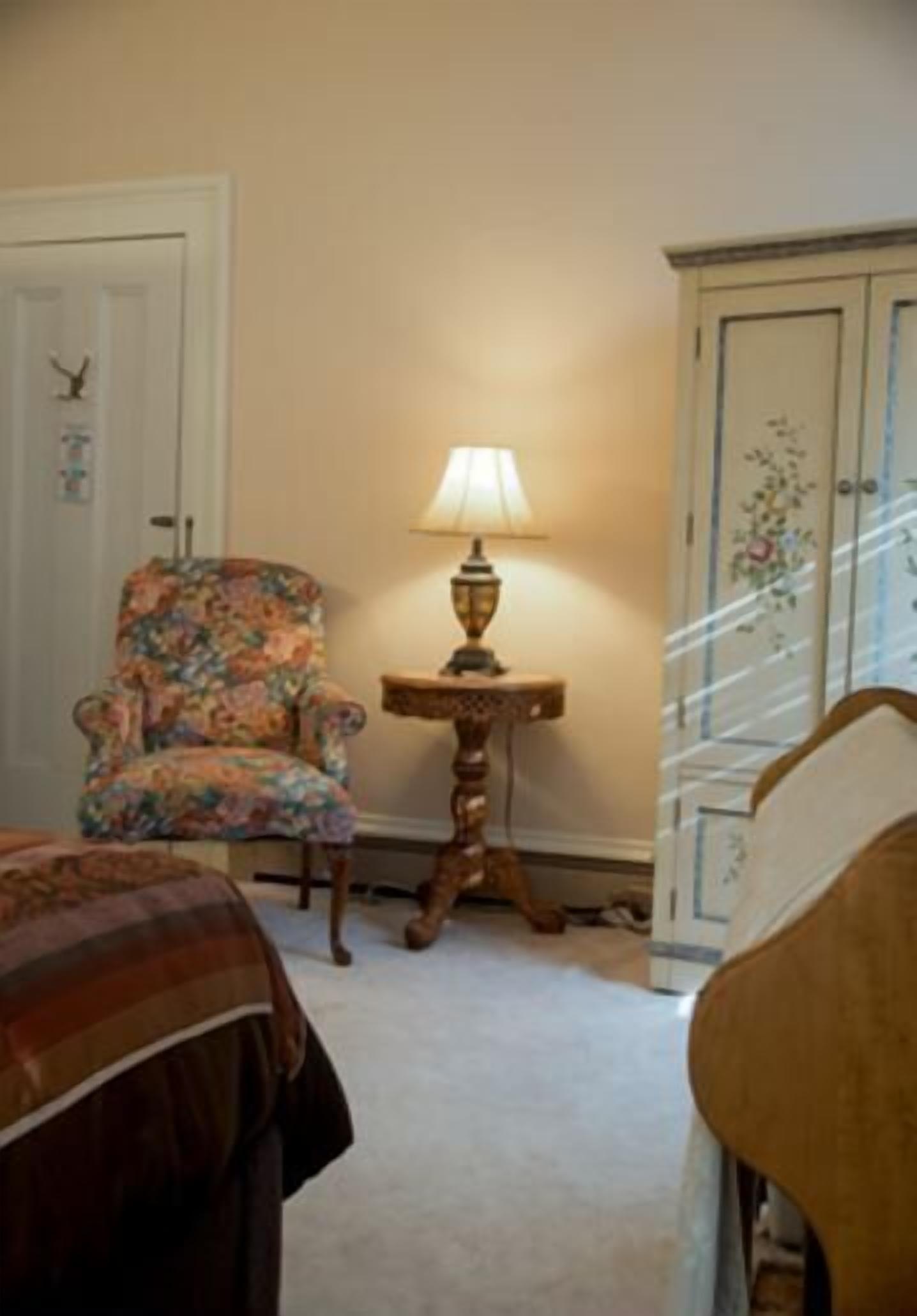 A room with a floral armchair, a table lamp, a wooden cabinet, and a bed.