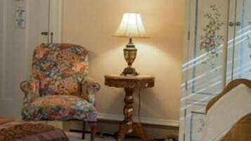 A room with a floral armchair, a table lamp, a wooden cabinet, and a bed.