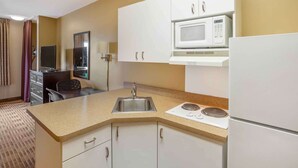 A compact hotel kitchen with white cabinets, a microwave, a refrigerator, a sink, and a small dining area with a chair and a flat-screen TV. - Extended Stay America Suites Meadowlands Rutherford (Rutherford)