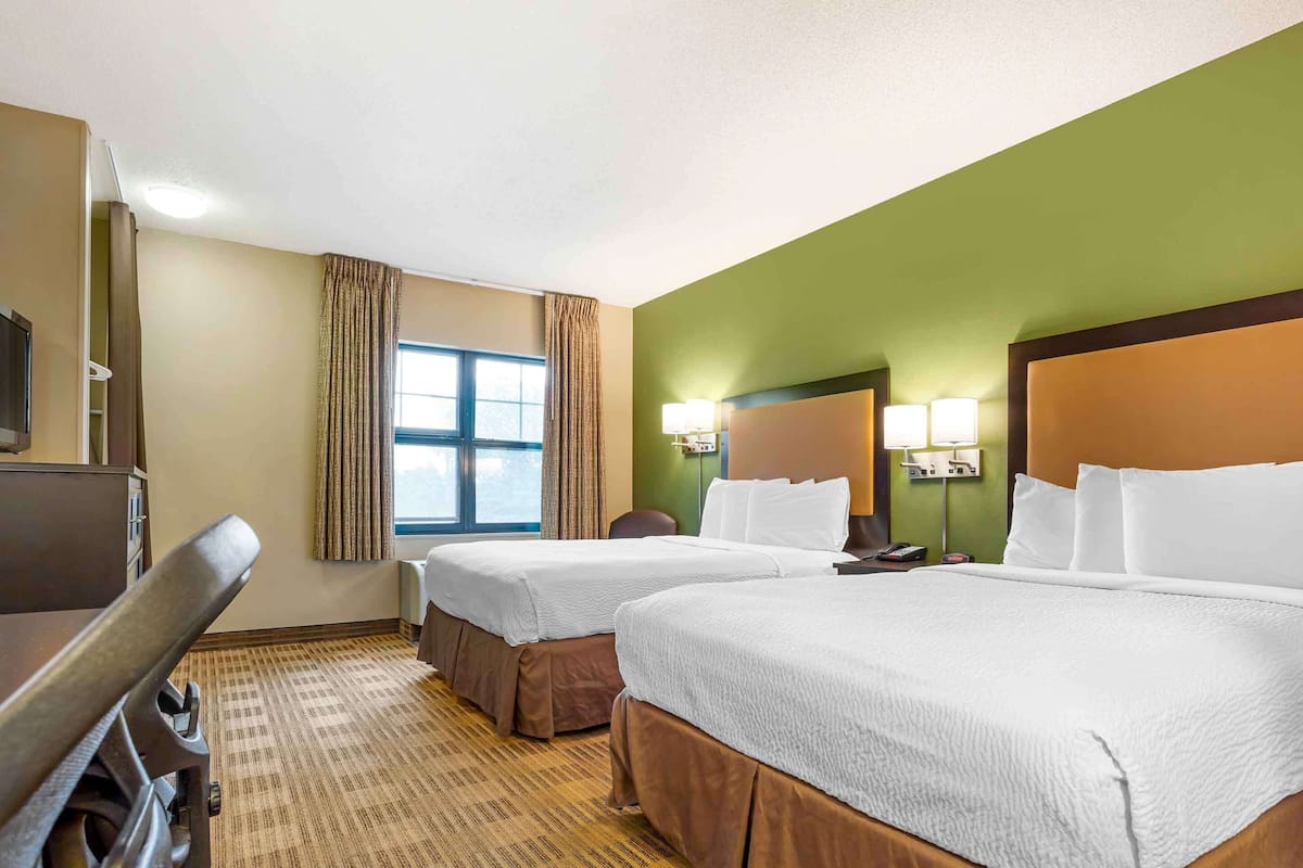A hotel room with two beds, a desk, a chair, a TV, and a window with curtains.