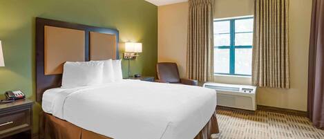 A hotel room with a large bed, a bedside table with a lamp, a chair, a window with curtains, and an air conditioning unit.