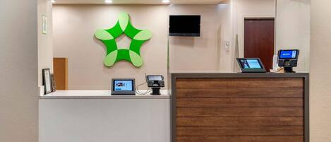 A reception area with a modern design, featuring a wooden reception desk, a green abstract wall decoration, a mounted television, and a digital kiosk.
