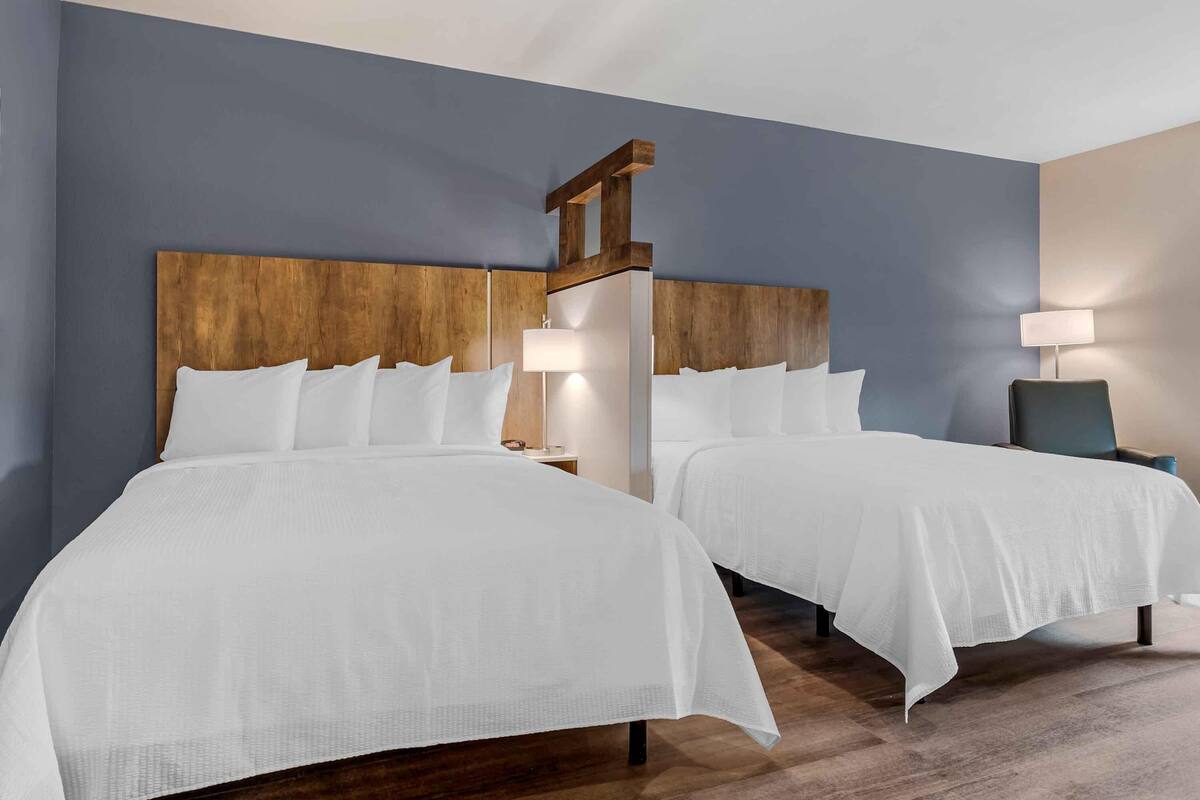 A hotel room with two beds, wooden headboards, white bedding, bedside lamps, and a chair.