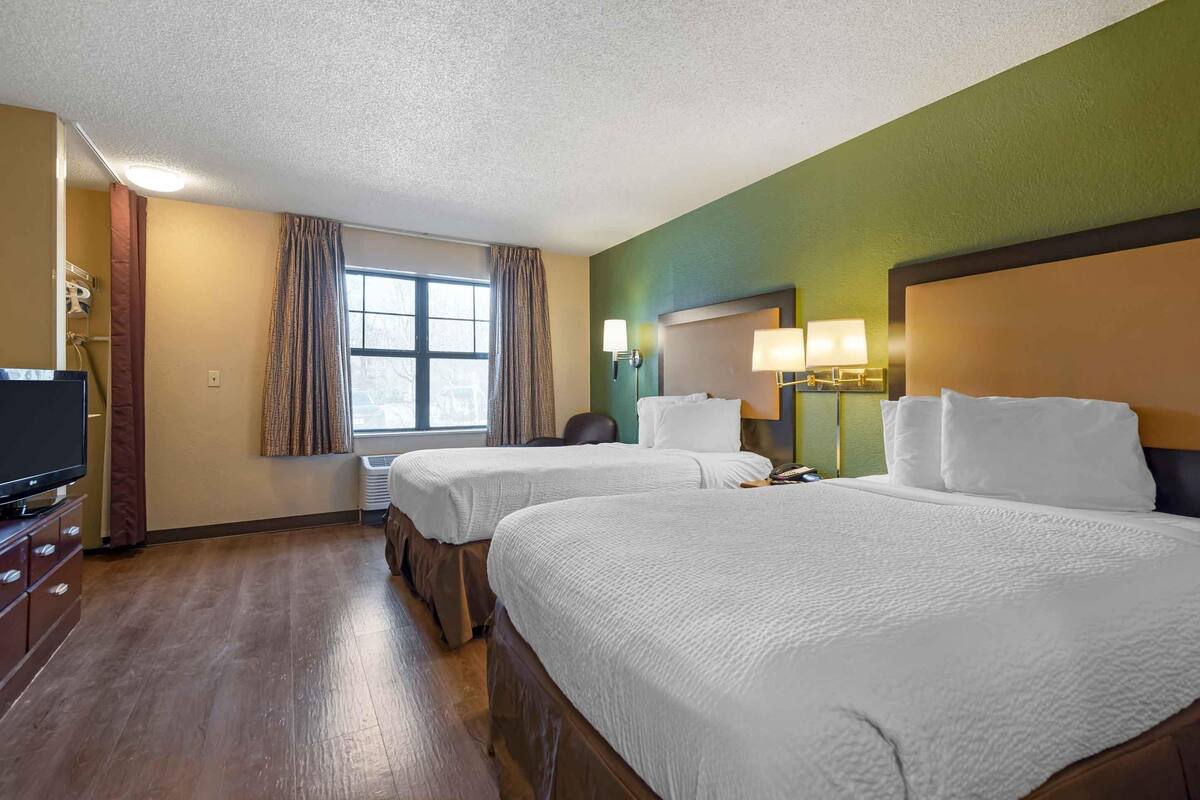 A hotel room with two beds, a television, a desk, and a window with curtains.