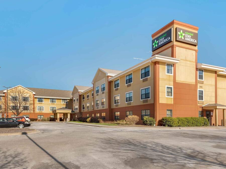 Un hotel de varios pisos con un letrero llamativo de Stay America, que cuenta con una entrada central y un estacionamiento con varios automóviles.