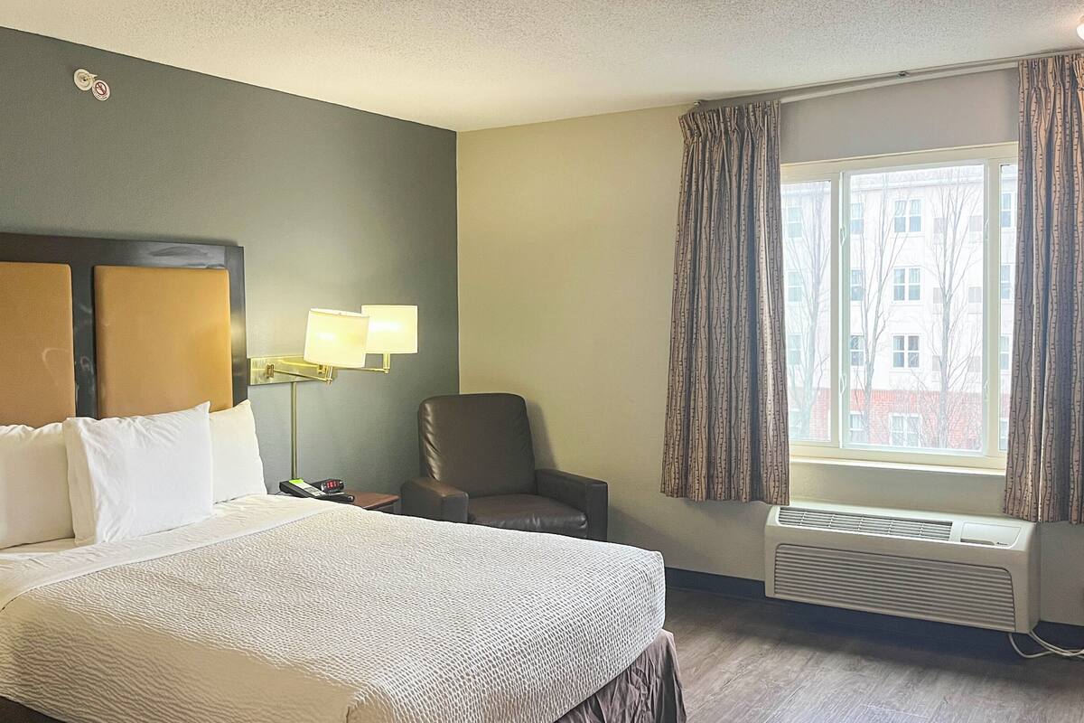 A hotel room with a bed, a chair, a desk with a lamp, and a window with curtains.
