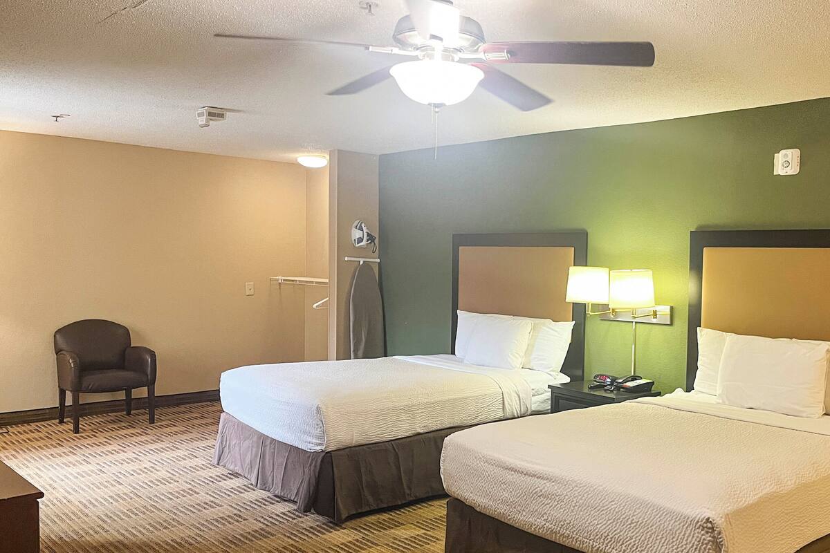 A hotel room with two beds, a ceiling fan, a nightstand with a lamp, and a chair.