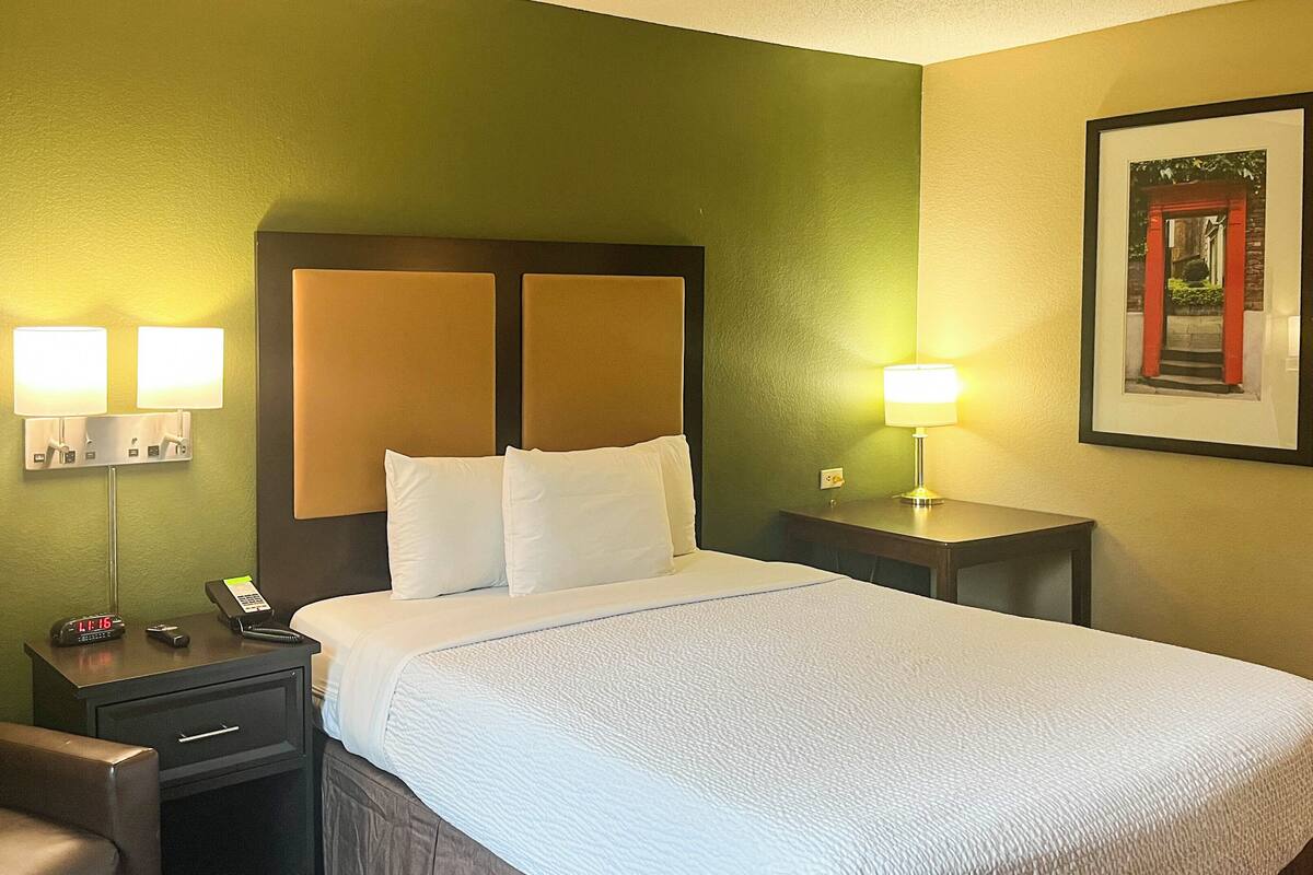 A hotel room with a bed, two bedside lamps, a chair, a nightstand, and a framed picture on the wall.