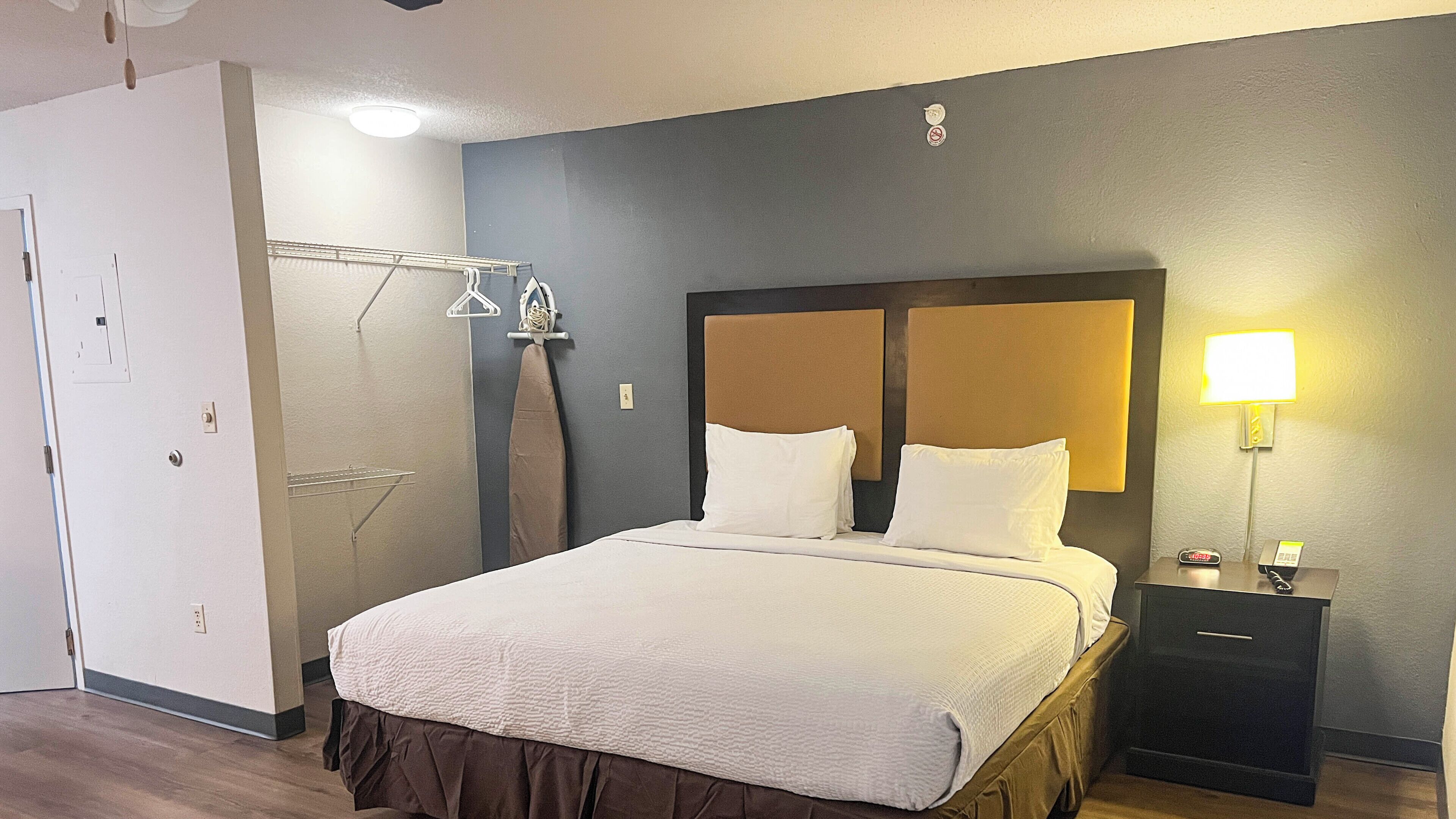 Una cama bien hecha con sábanas blancas, una mesita de noche con lámpara y un perchero para toallas en la pared.