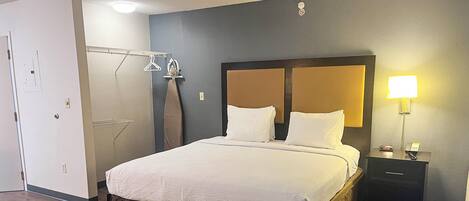 Una cama bien hecha con sábanas blancas, una mesita de noche con lámpara y un perchero para toallas en la pared.