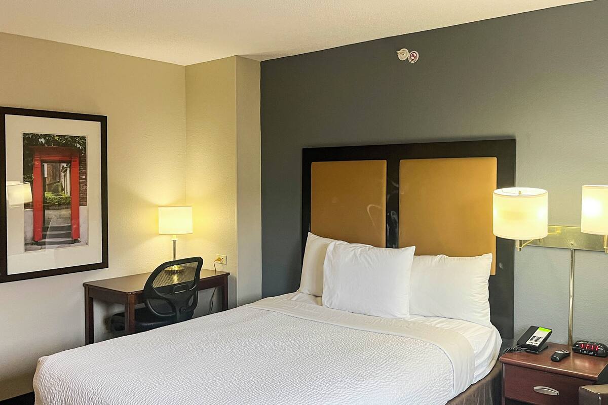 A hotel room with a bed, bedside lamps, a desk with a chair, a framed picture on the wall, and a nightstand with a phone and clock.