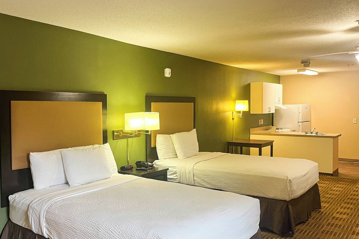 A hotel room with two beds, a desk, a lamp, a phone, and a kitchenette.