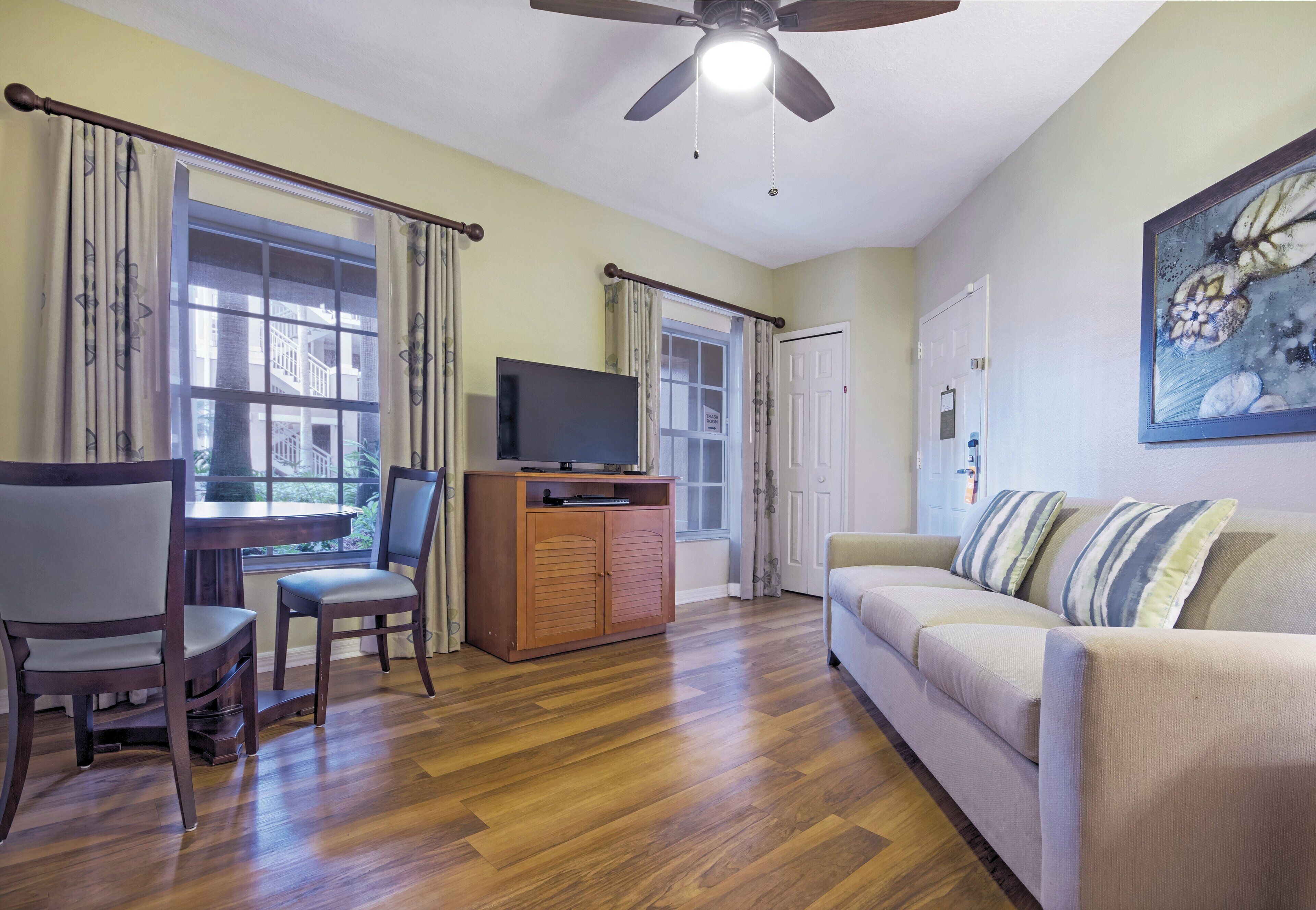 A living room with a wooden floor, a sofa, a TV on a wooden cabinet, and a ceiling fan.