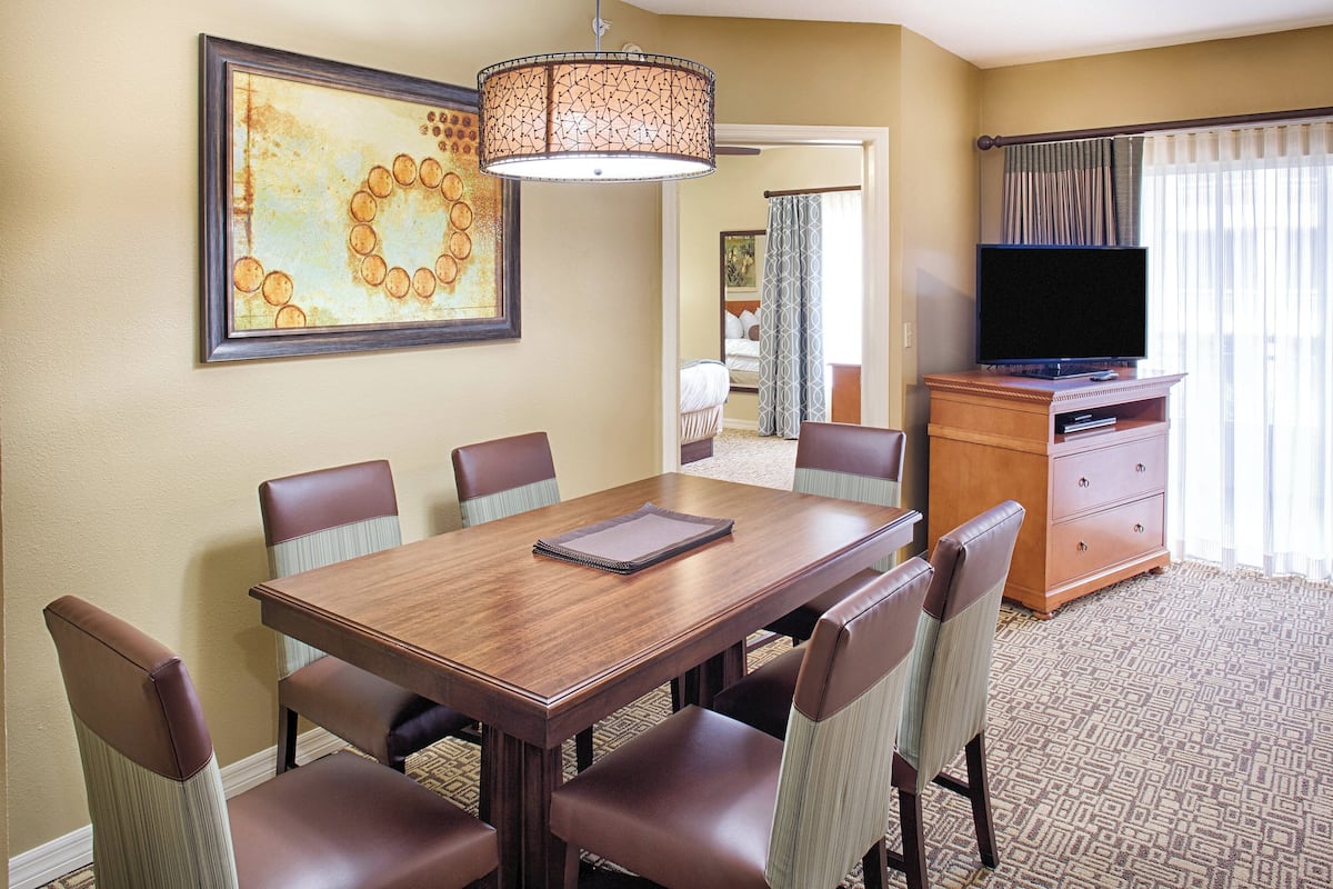 A hotel room with a dining table, six chairs, a television, and a painting on the wall.