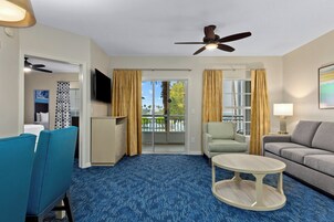 Habitación de hotel con alfombra azul, ventilador de techo, sofá, sillón, mesita y televisor.
