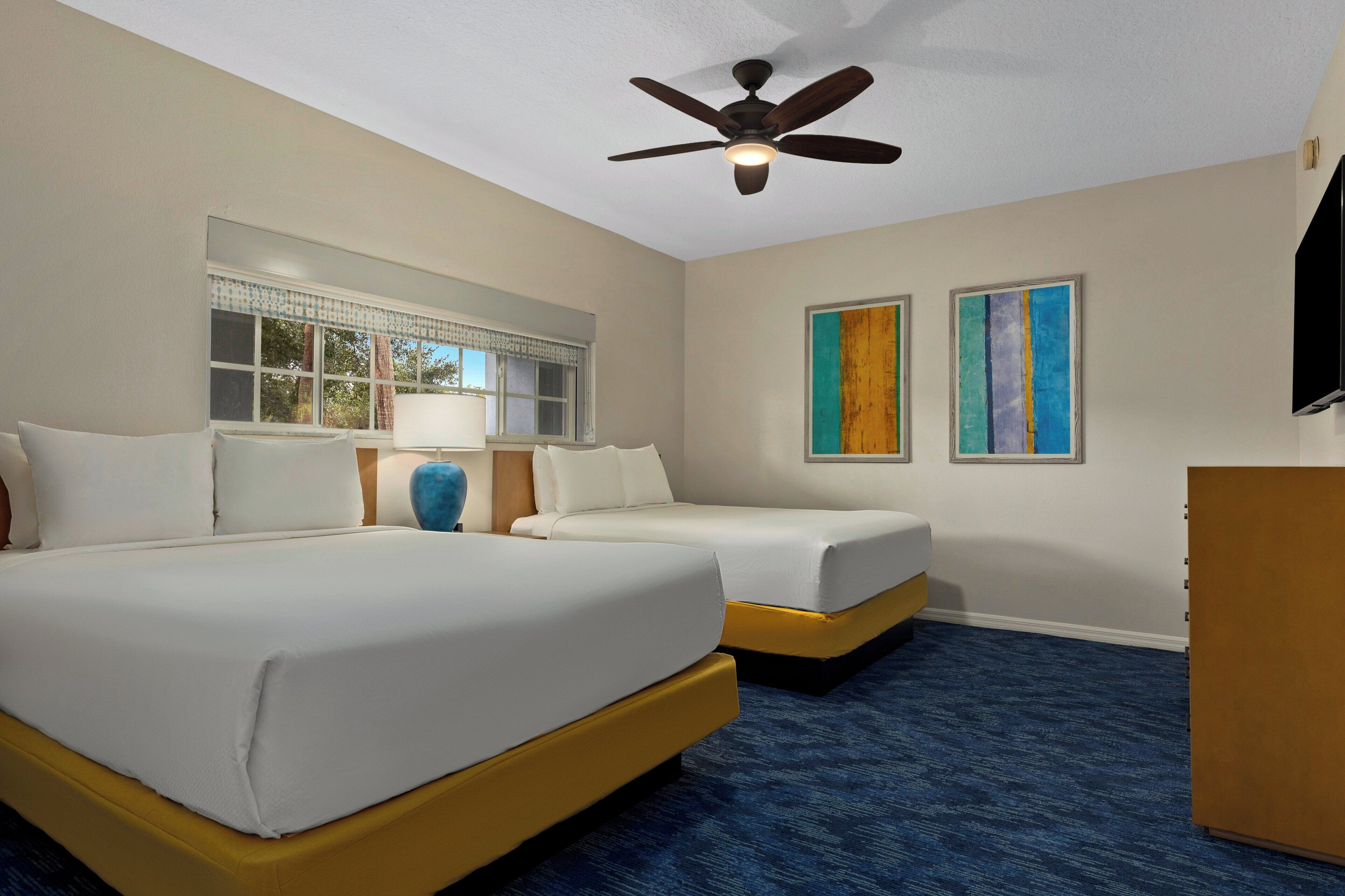 A hotel room with two beds, a ceiling fan, a window with blinds, and two framed artworks on the wall.