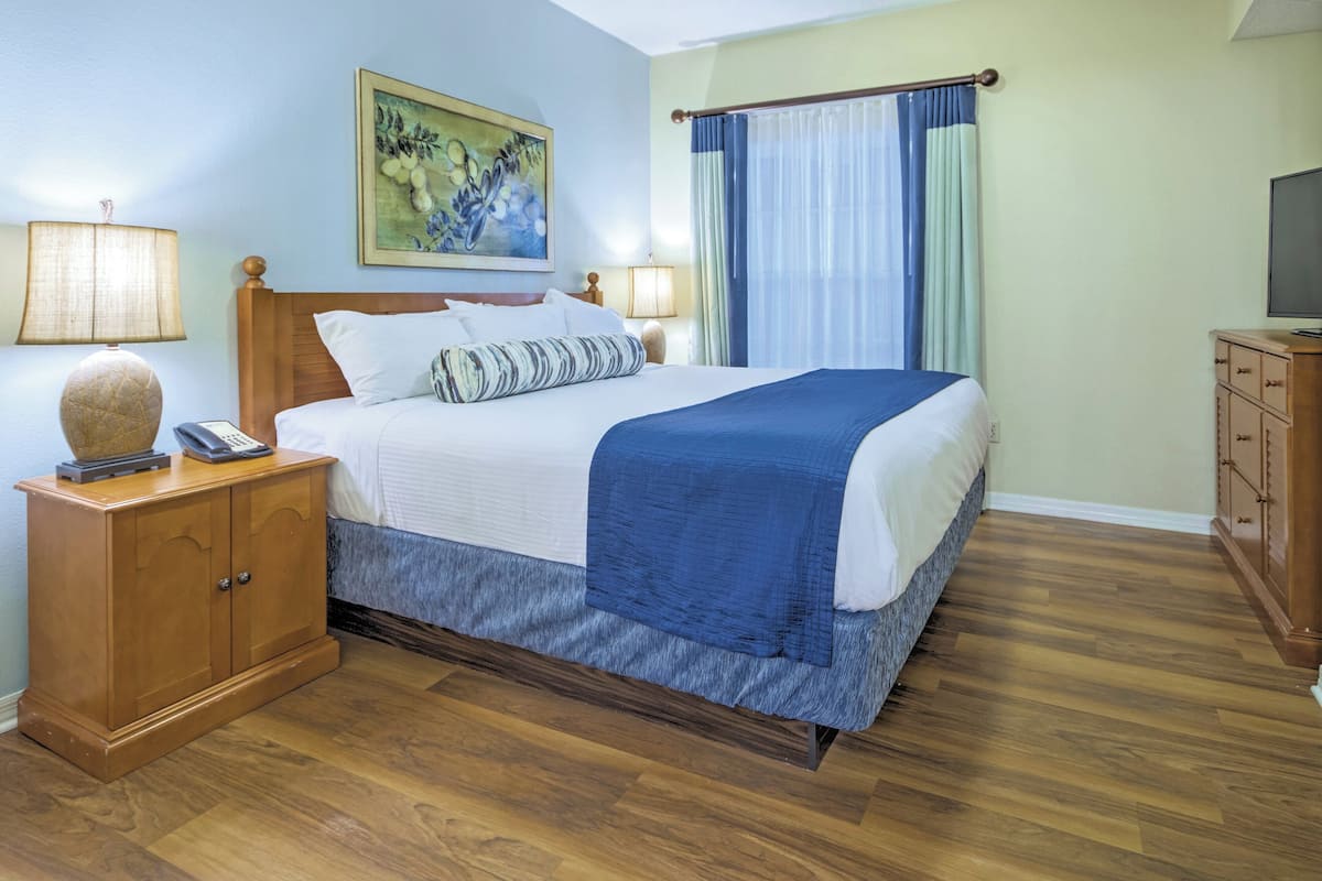 A hotel room with a wooden bed, a nightstand with a lamp, a wooden dresser, and a painting on the wall.