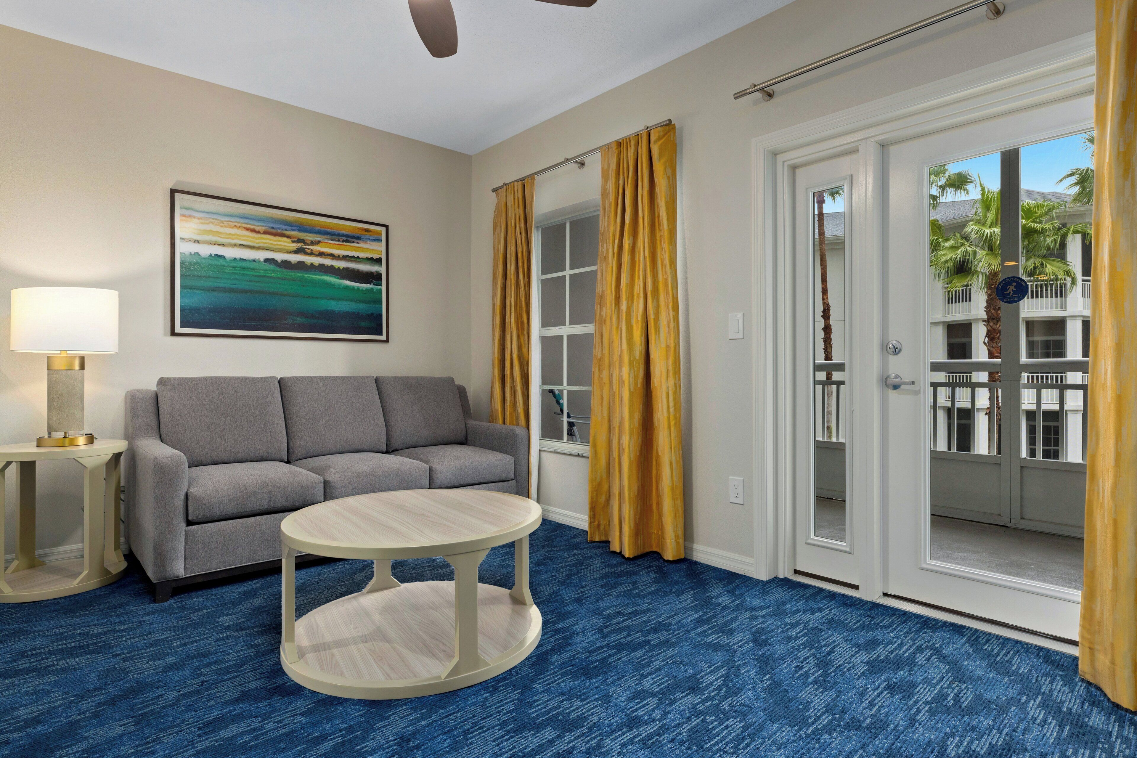 A hotel room with a gray sofa, a round table, a painting on the wall, and a view of a building with balconies through the door.