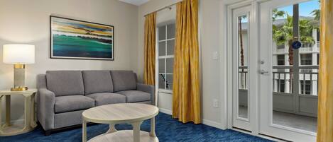 A hotel room with a gray sofa, a round table, a painting on the wall, and a view of a building with balconies through the door.