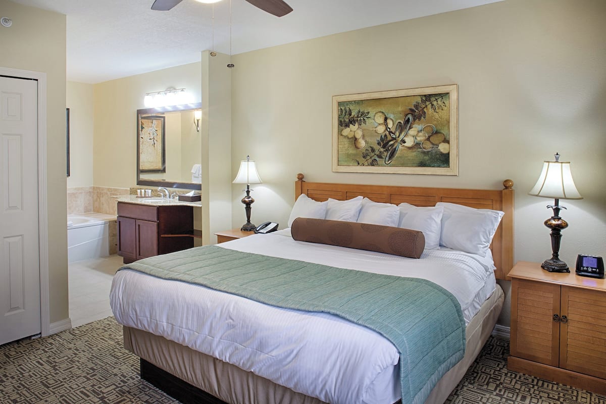A hotel room with a bed, a nightstand, a lamp, a mirror, a painting, and a bathroom with a sink and toilet.