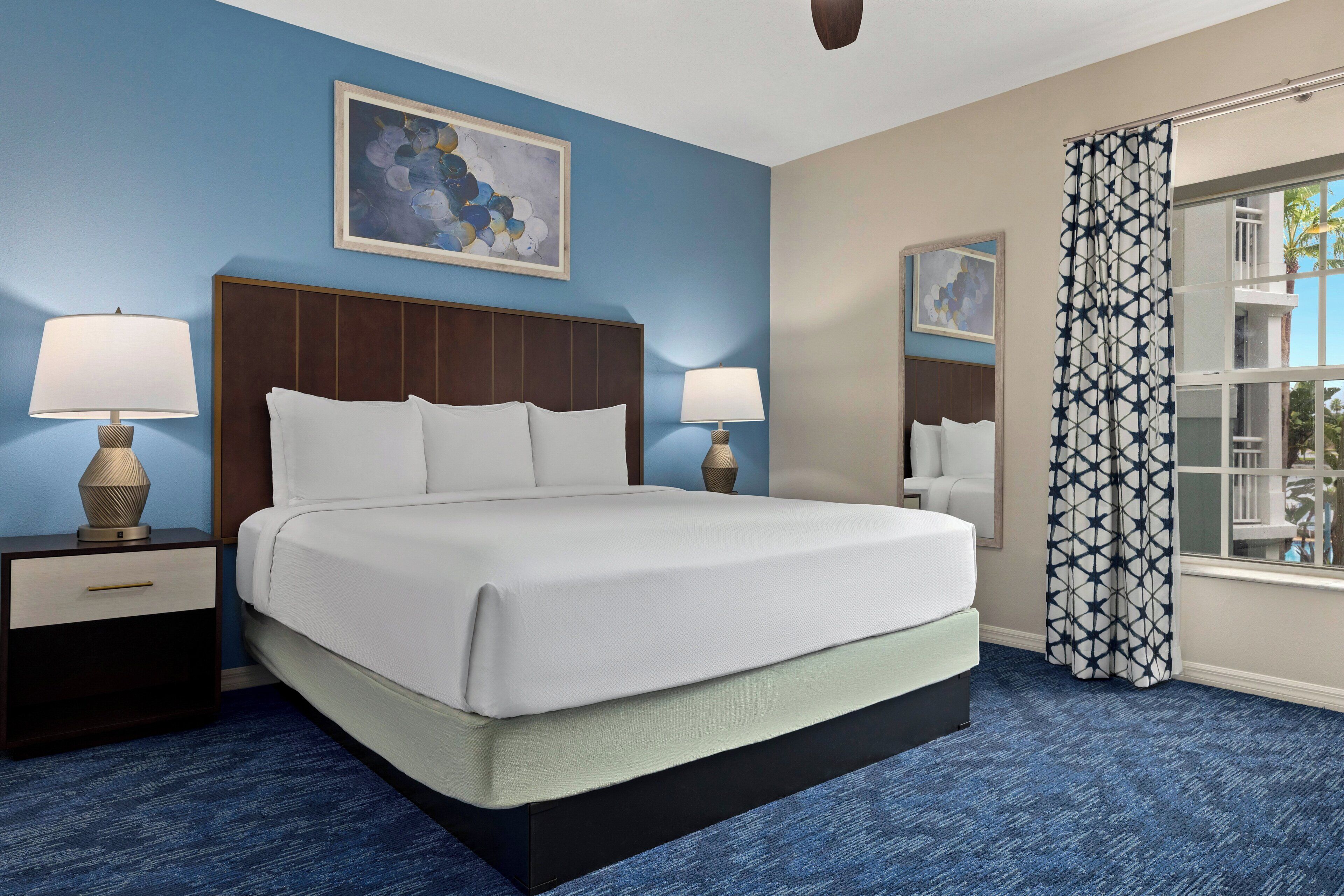 A hotel room with a large bed, two bedside tables with lamps, a ceiling fan, a window with a geometric curtain, and artwork on the walls.