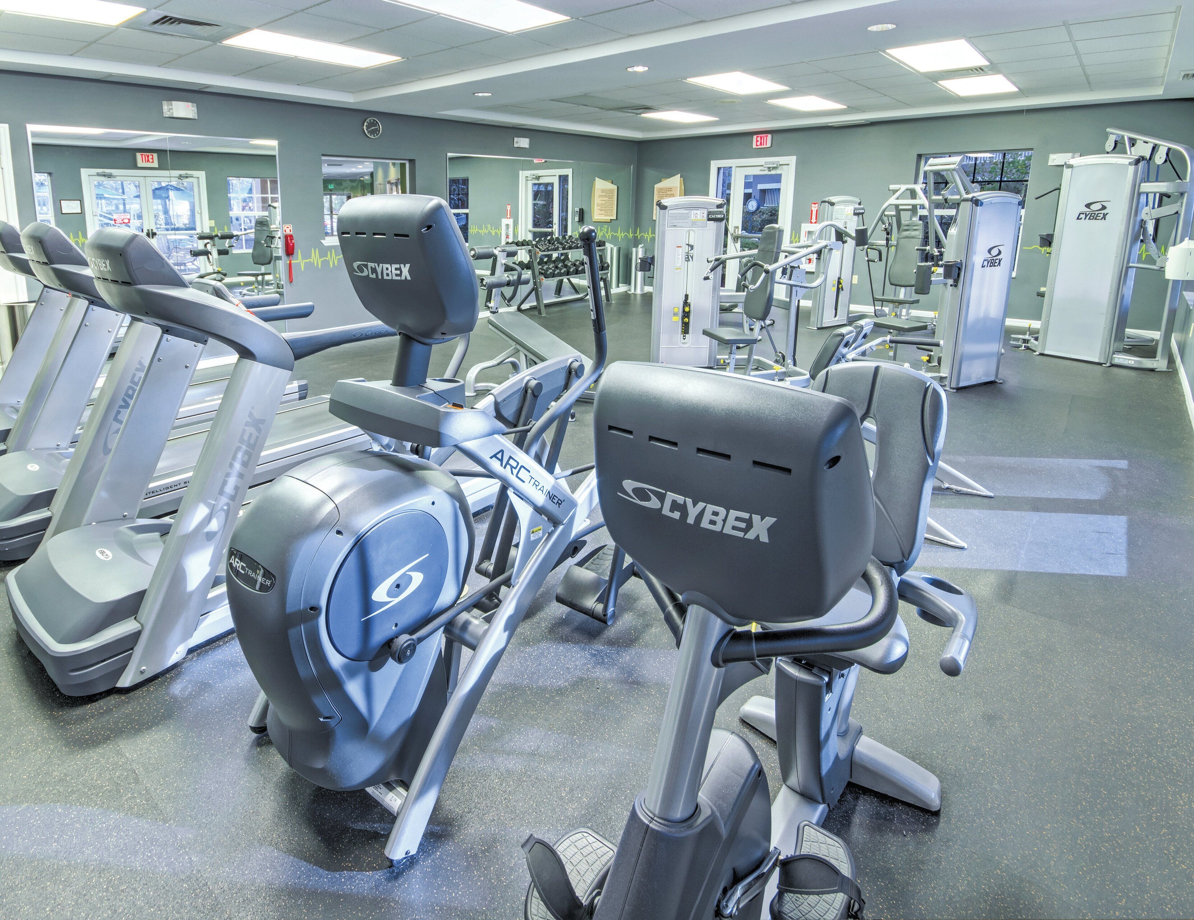 Uma academia com diversos equipamentos de exercício, incluindo esteiras e bicicletas ergométricas, chão cinzento e paredes com espelhos.