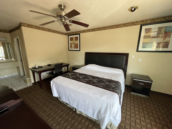 A hotel room with a bed, a desk, a chair, a nightstand, a ceiling fan, and a framed picture on the wall.