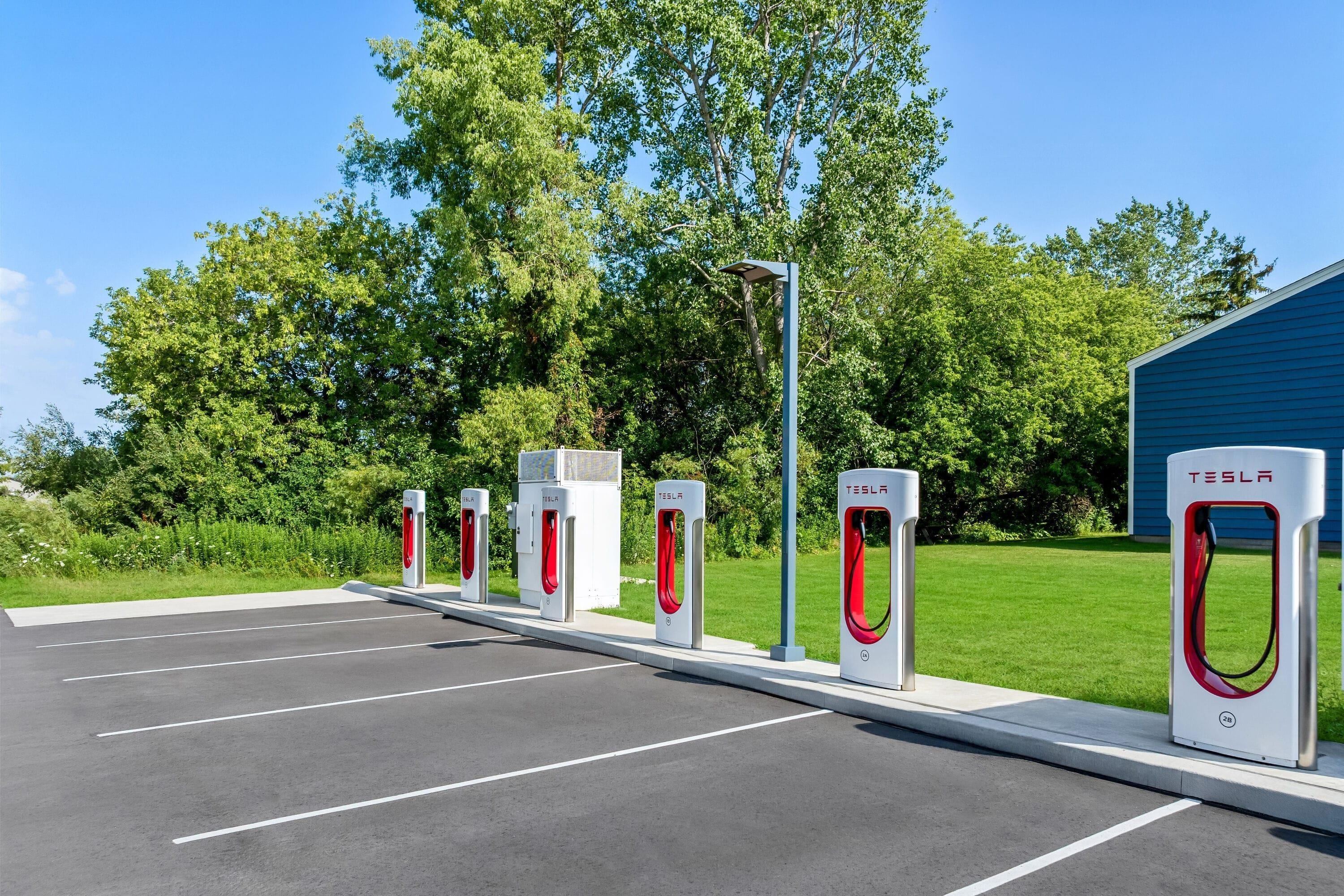 Una fila de estaciones de carga para vehículos eléctricos Tesla en un estacionamiento, con un edificio al fondo.